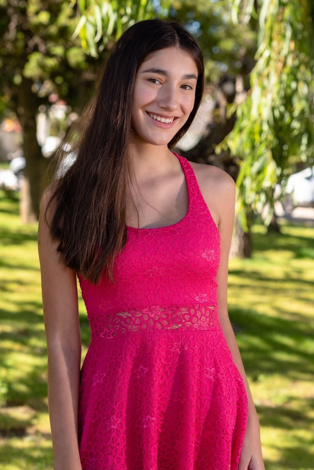 Mujer sonriendo con un vestido rosa, parada afuera con árboles en el fondo.