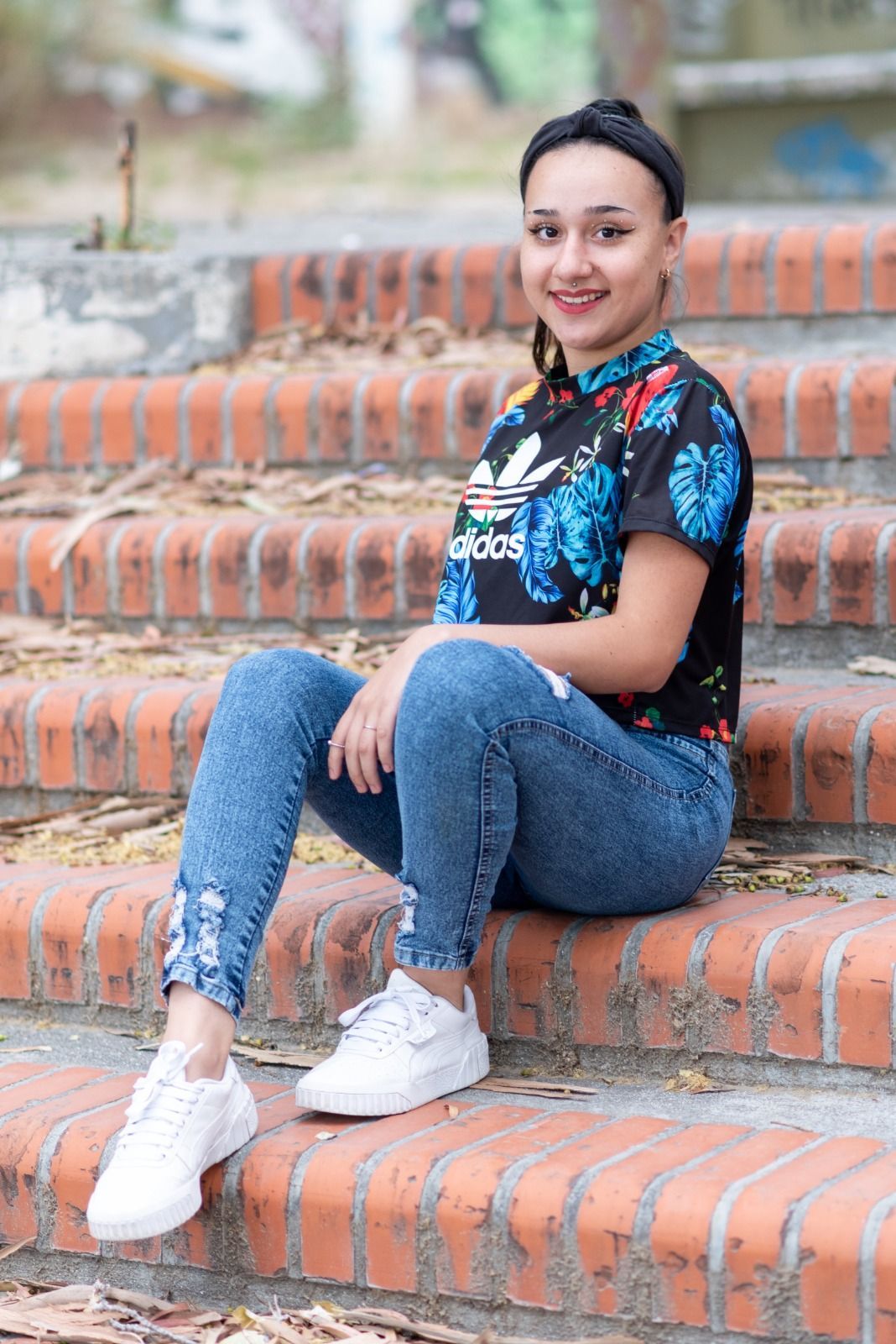 Una mujer con vaqueros y una blusa floreada se sienta en unos escalones de ladrillo, sonriendo.