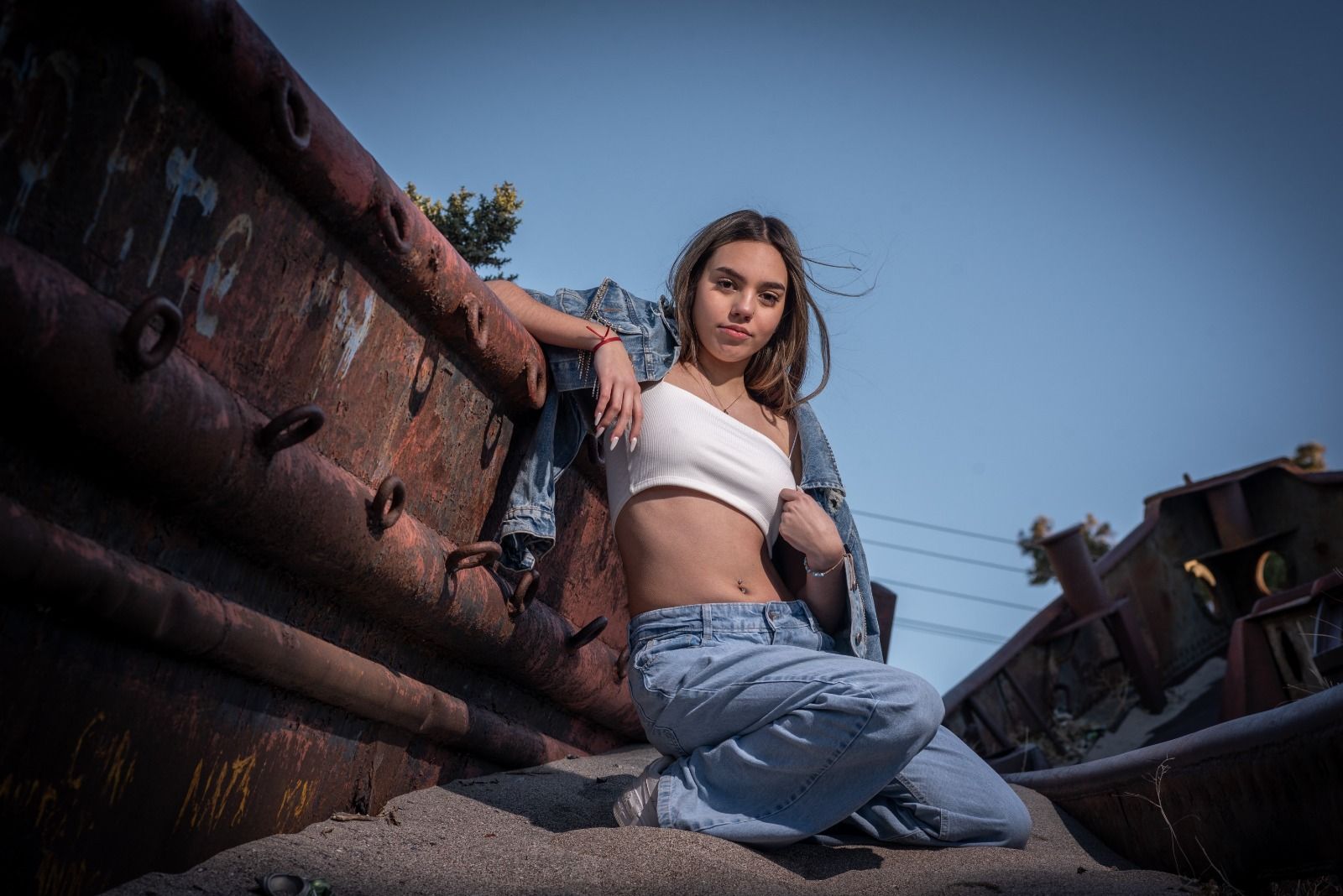 Mujer con top blanco y jeans posa cerca de estructuras de metal oxidadas.