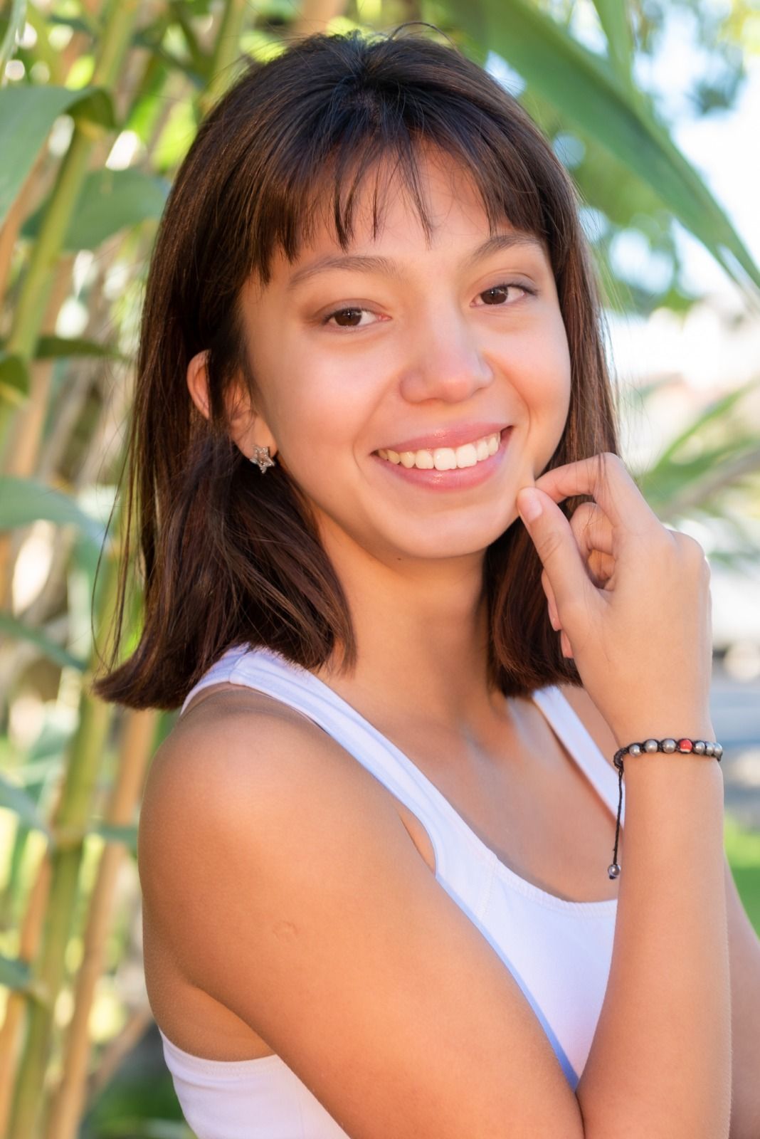Mujer sonriendo, tocándose la barbilla, vestida con un top blanco, al aire libre con vegetación.