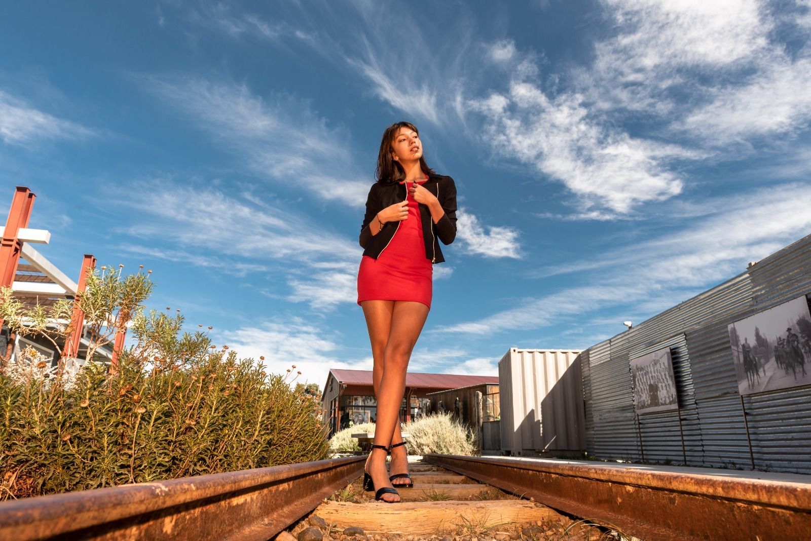 Una mujer con un vestido rojo y una chaqueta negra camina por las vías del tren oxidadas bajo un cielo azul.