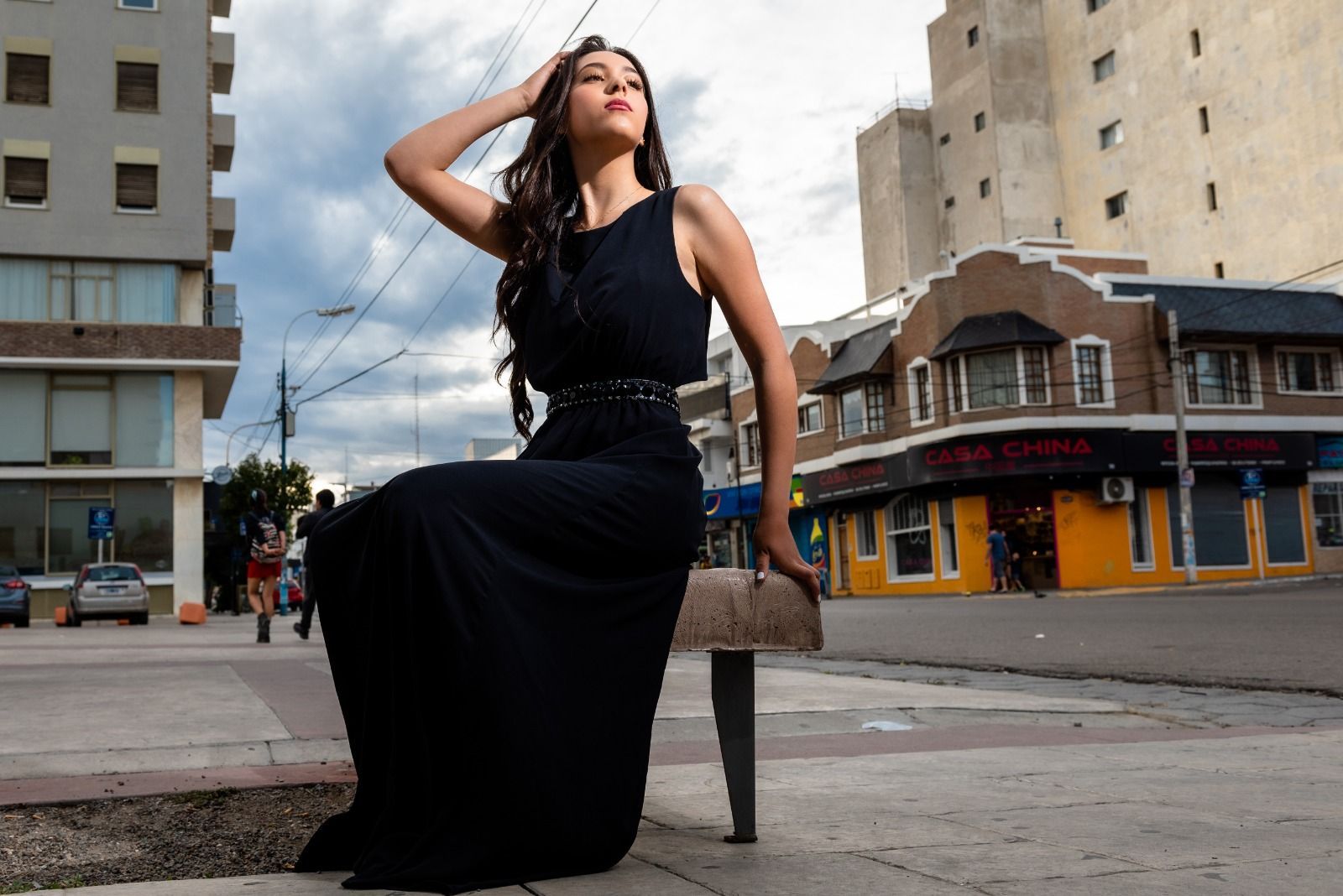 Mujer vestida de negro posa al aire libre, con la mano en la cabeza y edificios al fondo.