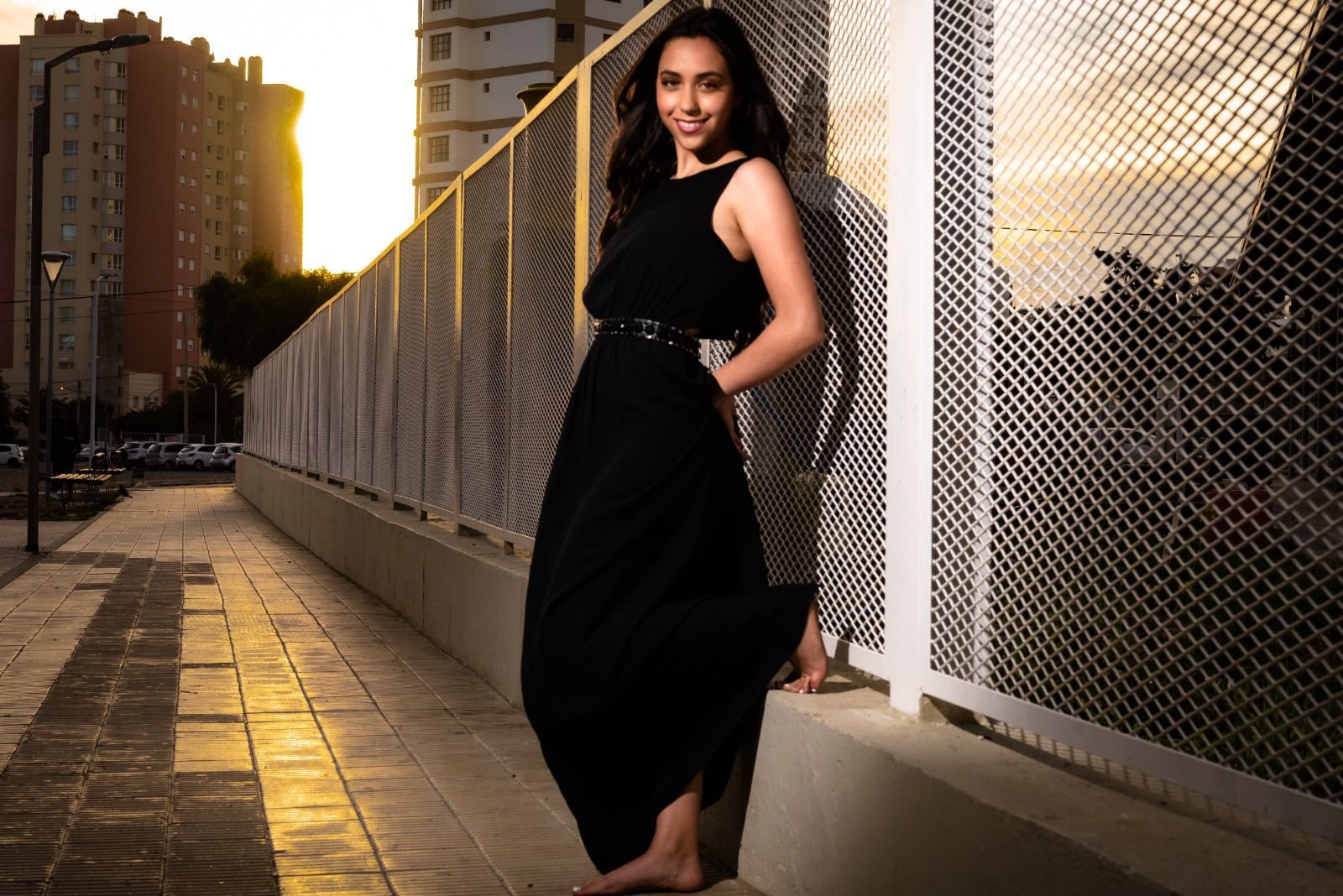 Una mujer vestida de negro se apoya contra una valla al aire libre al atardecer.