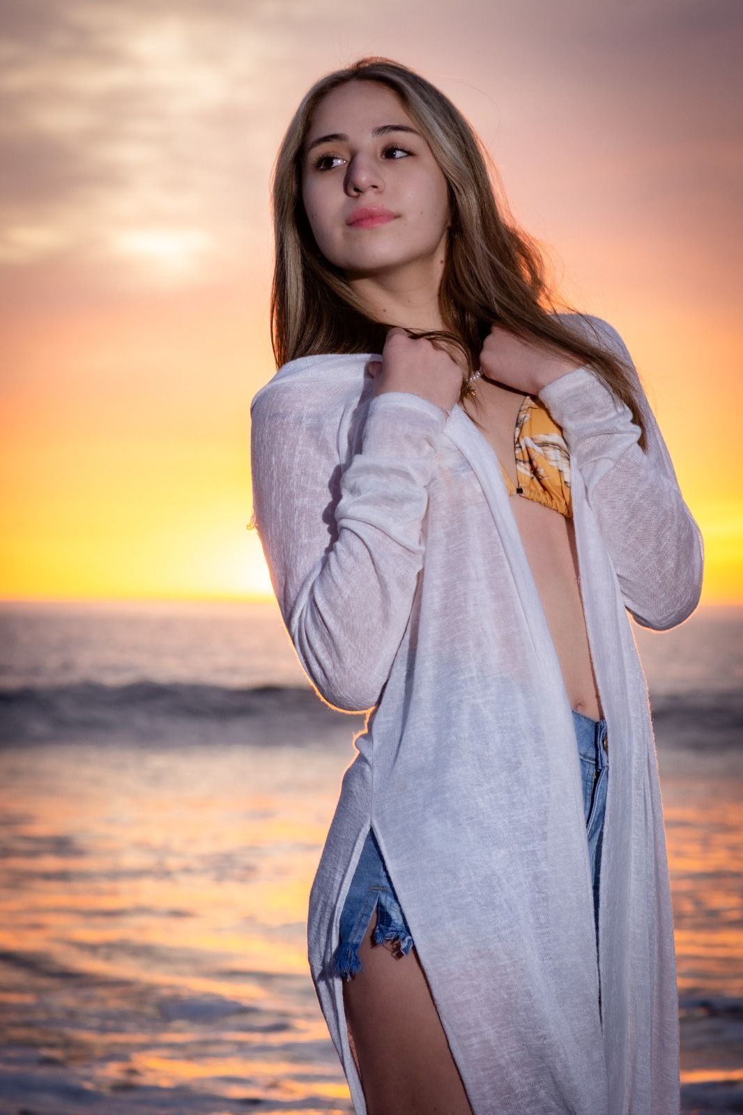 Mujer con cárdigan blanco y pantalones cortos de mezclilla en la playa al atardecer.