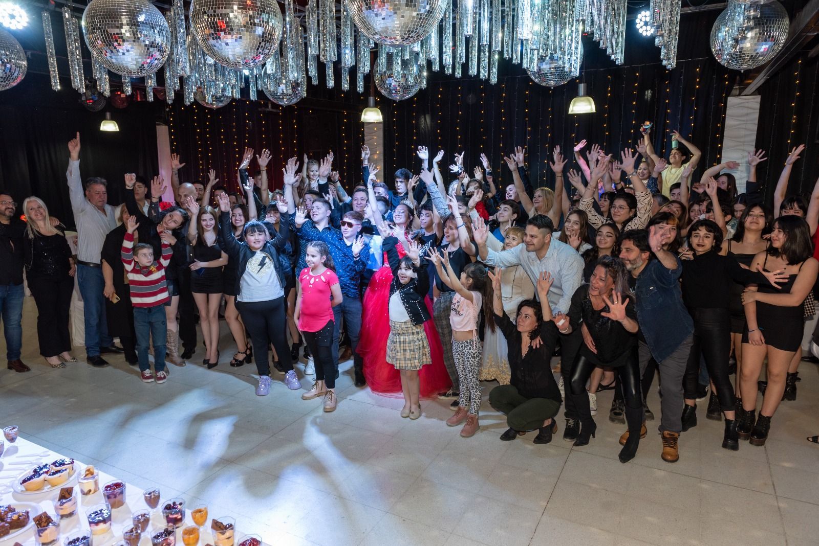 Gran grupo de personas con los brazos en alto, celebrando bajo bolas de discoteca y luces.