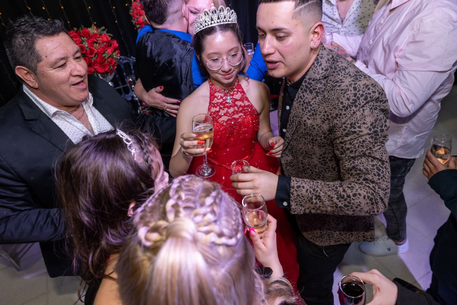 Gente brindando con champán; una persona joven con una tiara y un vestido rojo se encuentra en medio de una celebración.
