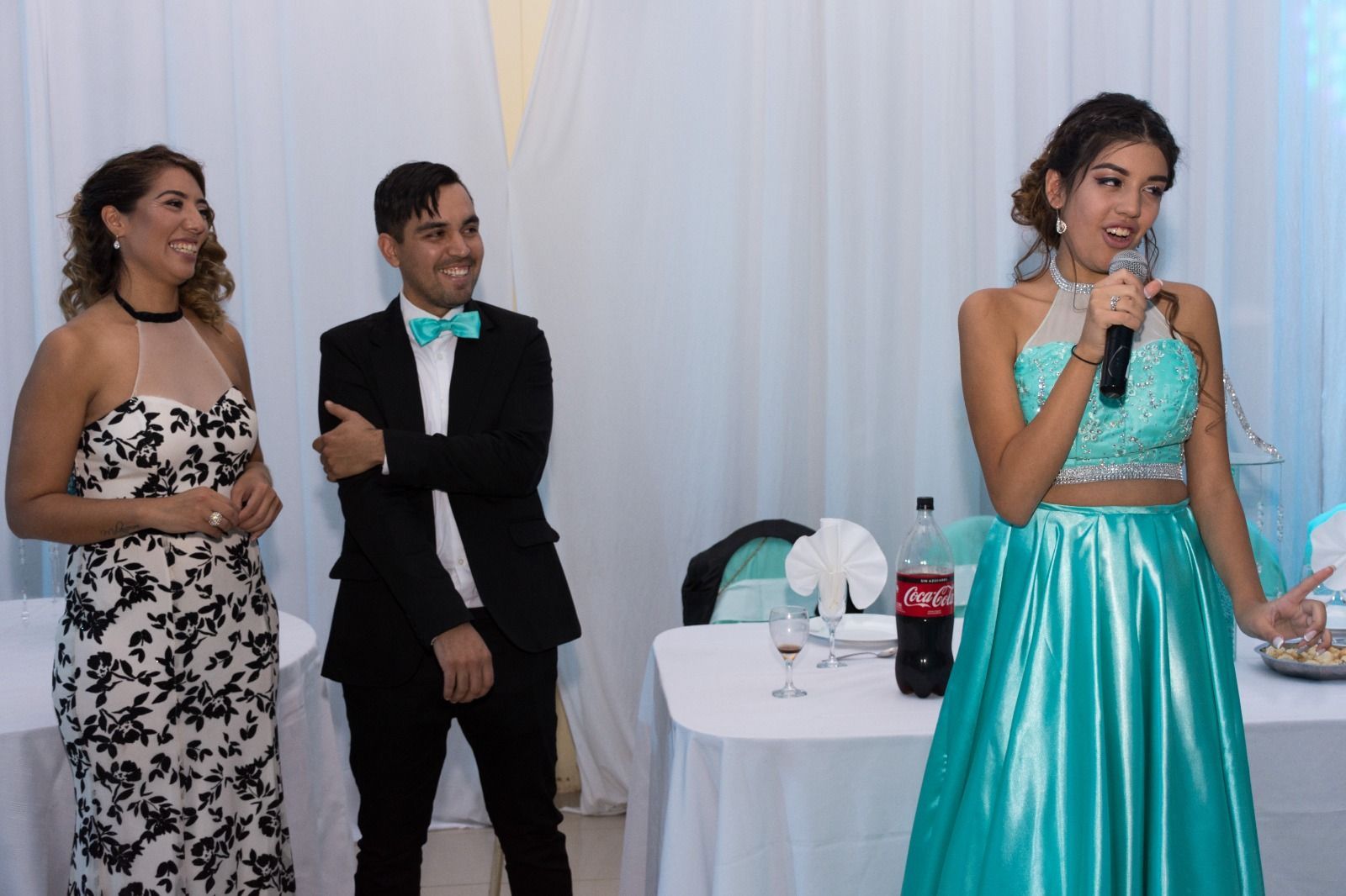 Mujer vestida de verde azulado hablando, dos personas escuchan en una fiesta, con manteles y decoraciones blancas.