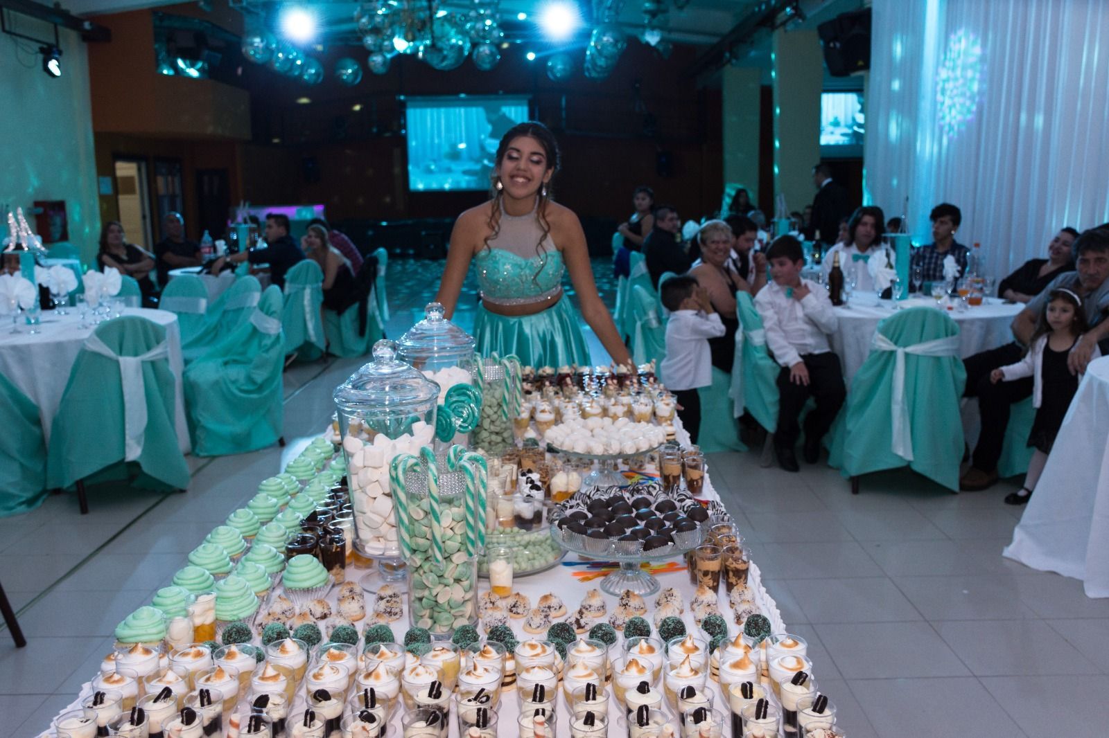 Una joven vestida de color turquesa se encuentra detrás de una mesa de postres en un evento, rodeada de invitados.
