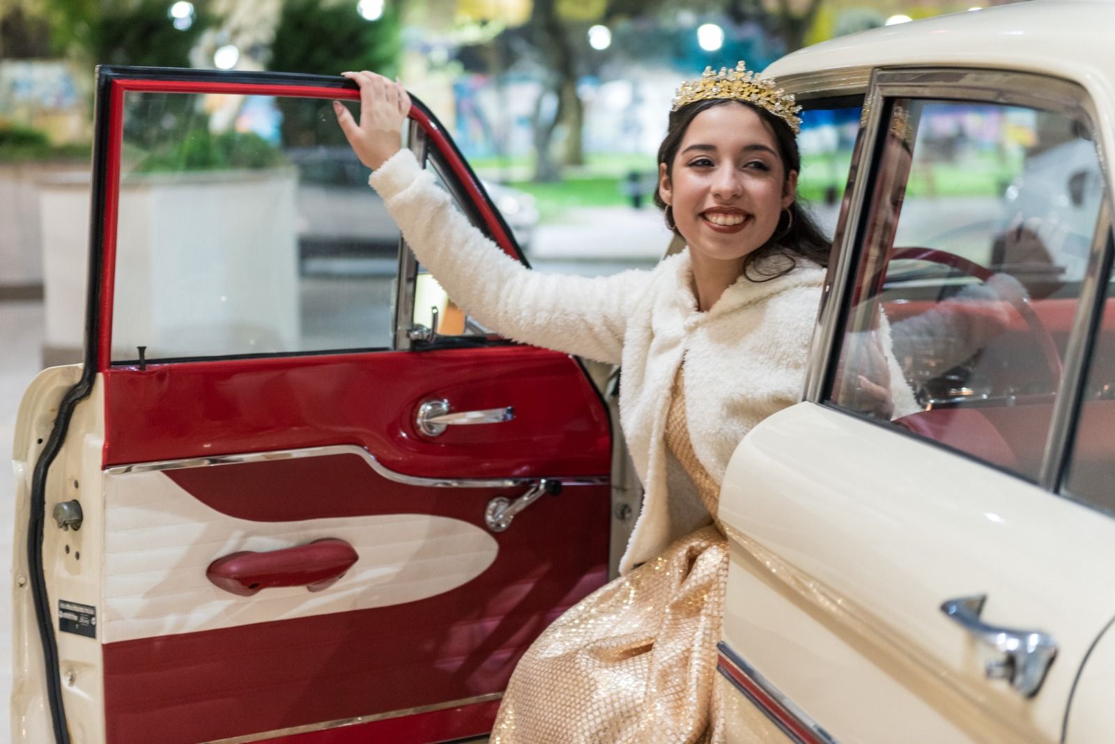 Una mujer con vestido dorado y corona sale de un automóvil clásico rojo y blanco, sonriendo.