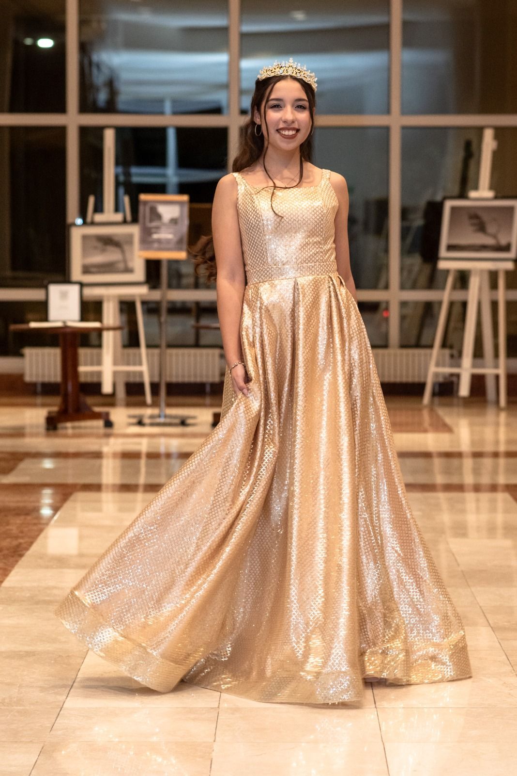 Mujer con un vestido dorado brillante y una corona, posando frente a ventanas con exhibiciones de arte.