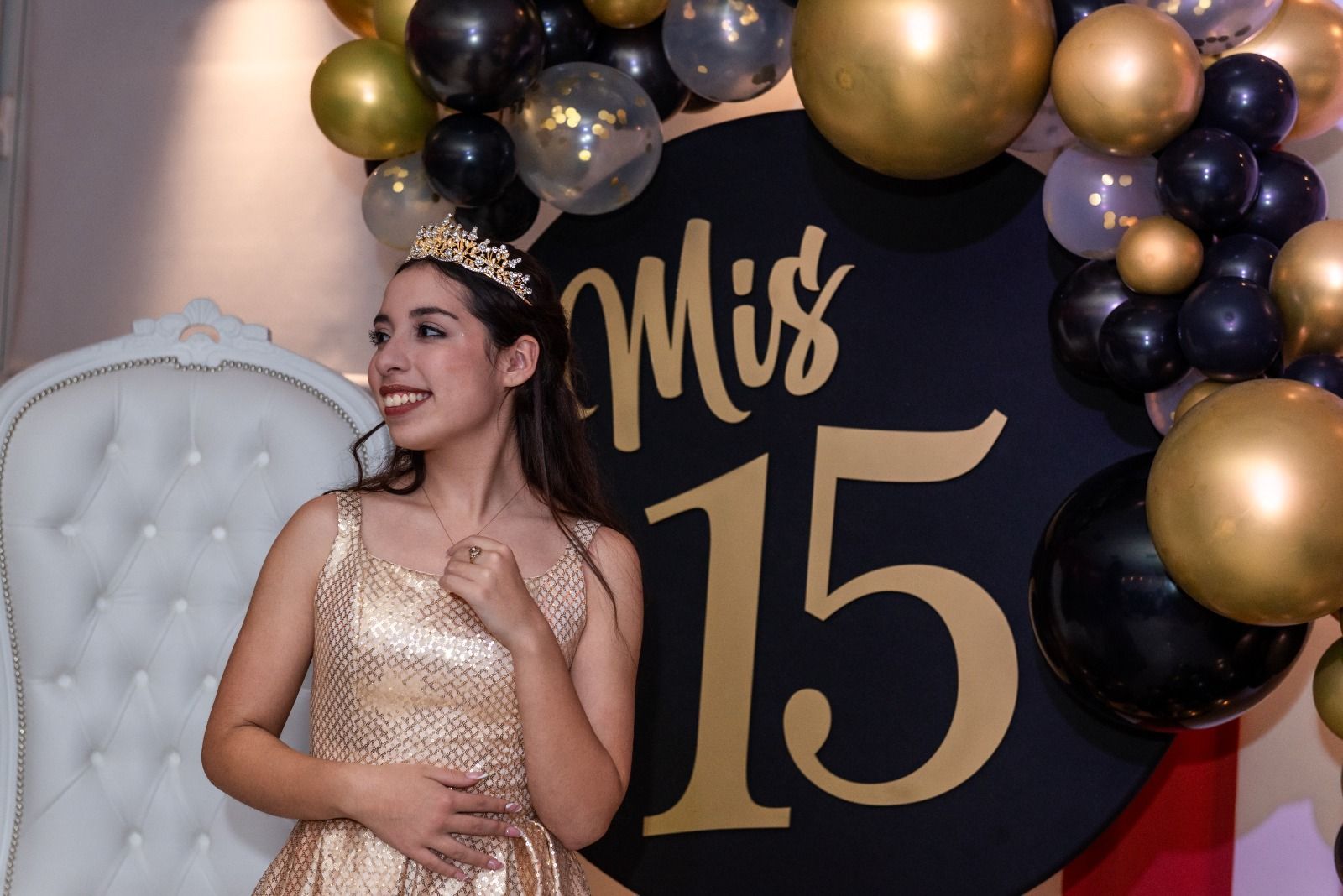 Mujer con vestido dorado y tiara en una celebración de quinceañera, posando cerca de un cartel de 