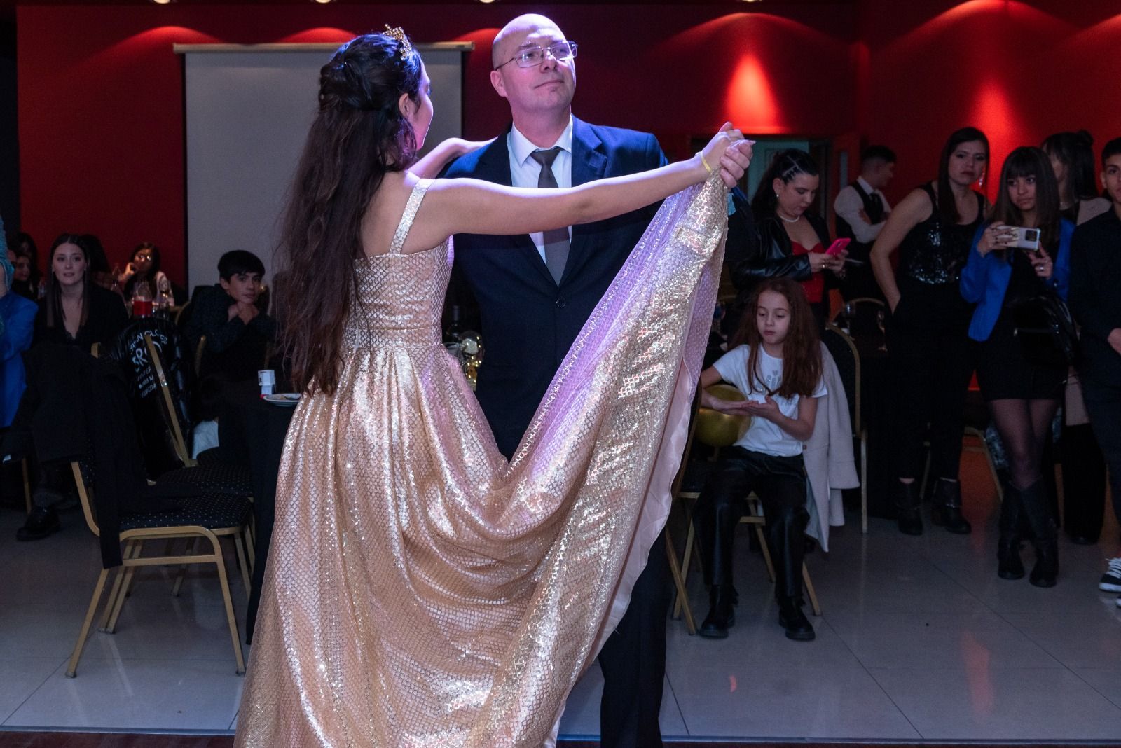 Un hombre y una mujer bailan, sosteniendo el vestido dorado de la mujer, en un evento. Otros observan.