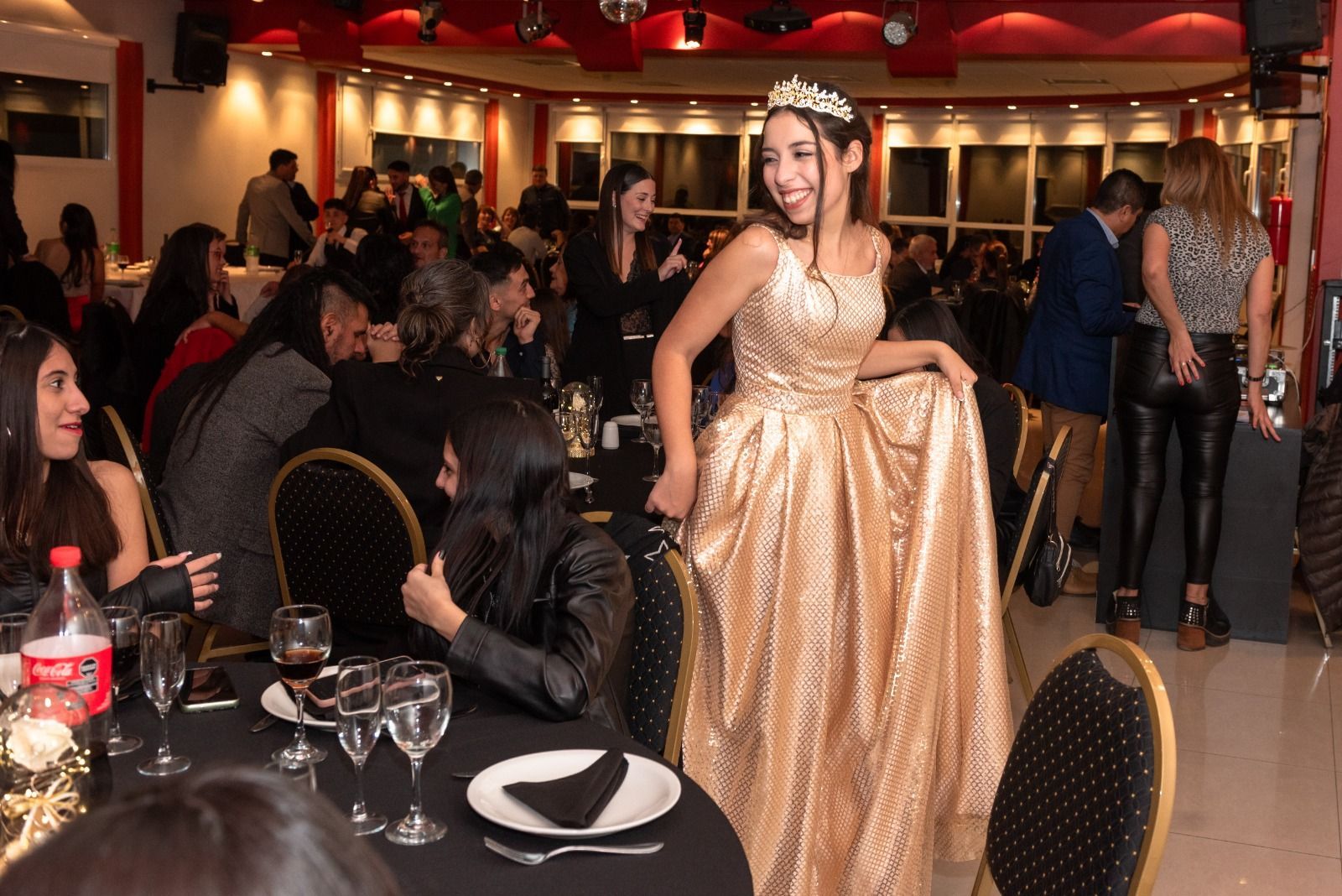Mujer con vestido dorado y tiara, sonriendo en una fiesta. Se ven mesas, invitados y una sala decorada con detalles en rojo.