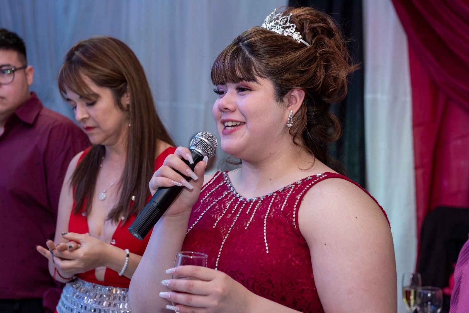 Una mujer con un vestido rojo y tiara habla por el micrófono en una celebración, mientras otras personas están cerca.