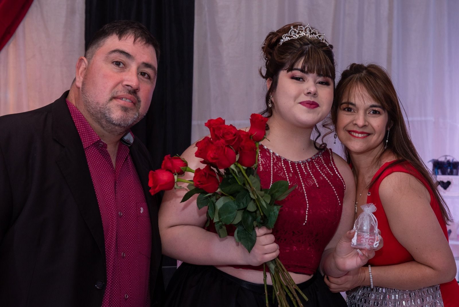 Familia posando en un evento. Un joven con vestido rojo sostiene rosas, flanqueado por un hombre y una mujer.