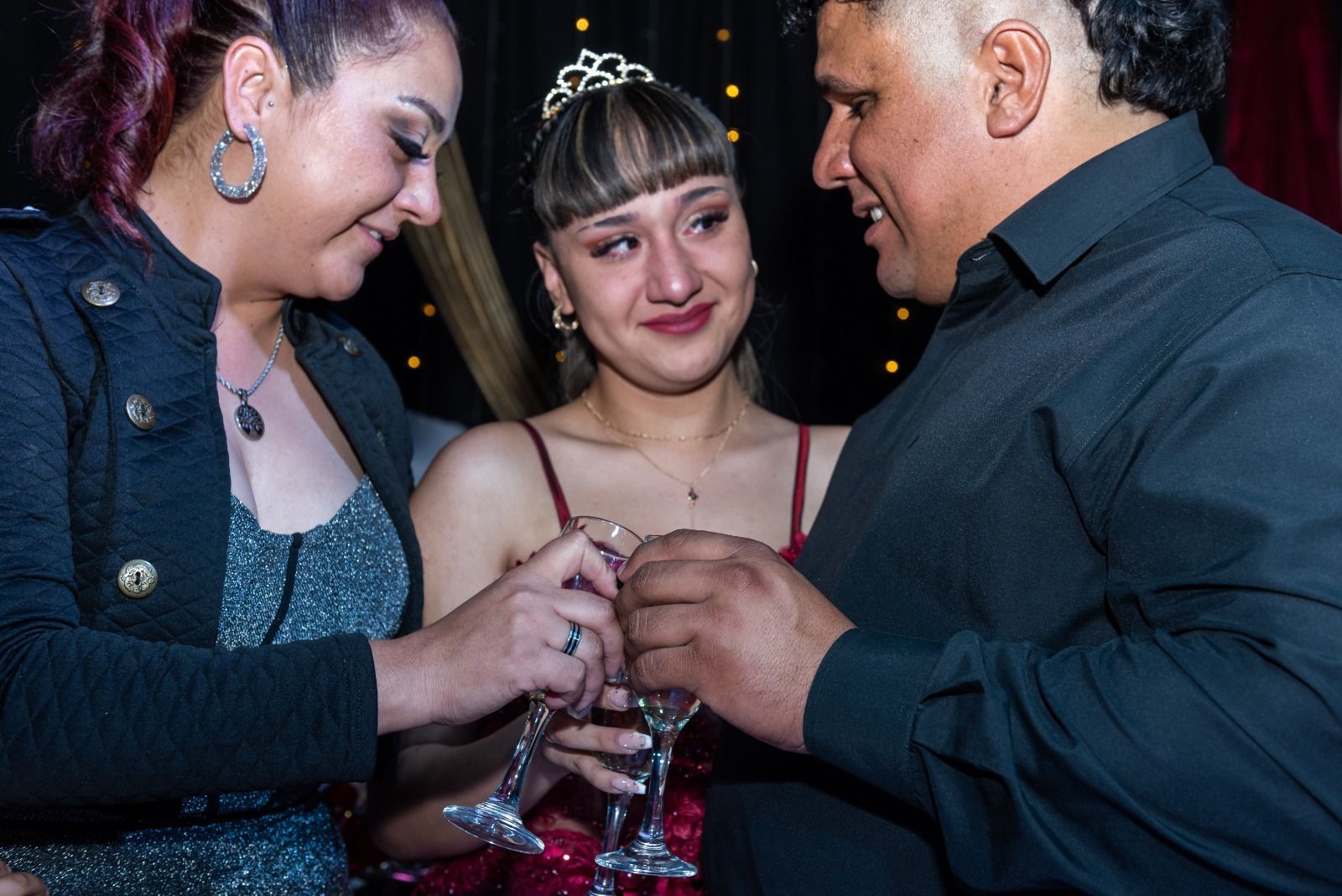 Familia brindando con champán. Una niña con vestido rojo, con lágrimas en los ojos, está rodeada de sus padres en una fiesta.