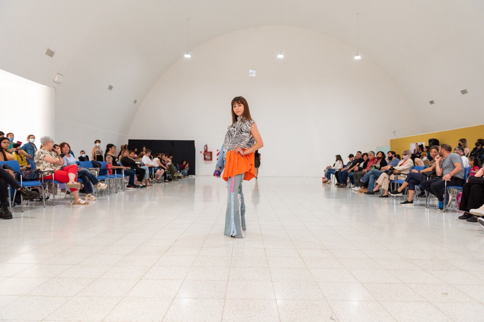 Una modelo camina por una pasarela en un espacio de paredes blancas; los espectadores la observan desde filas de sillas.