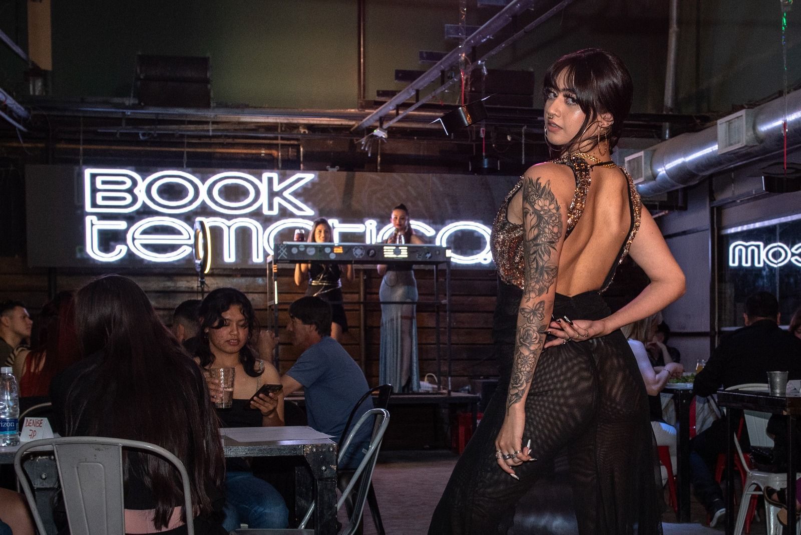 Una mujer vestida de negro posa en un local; detrás de ella hay un cartel con las palabras 