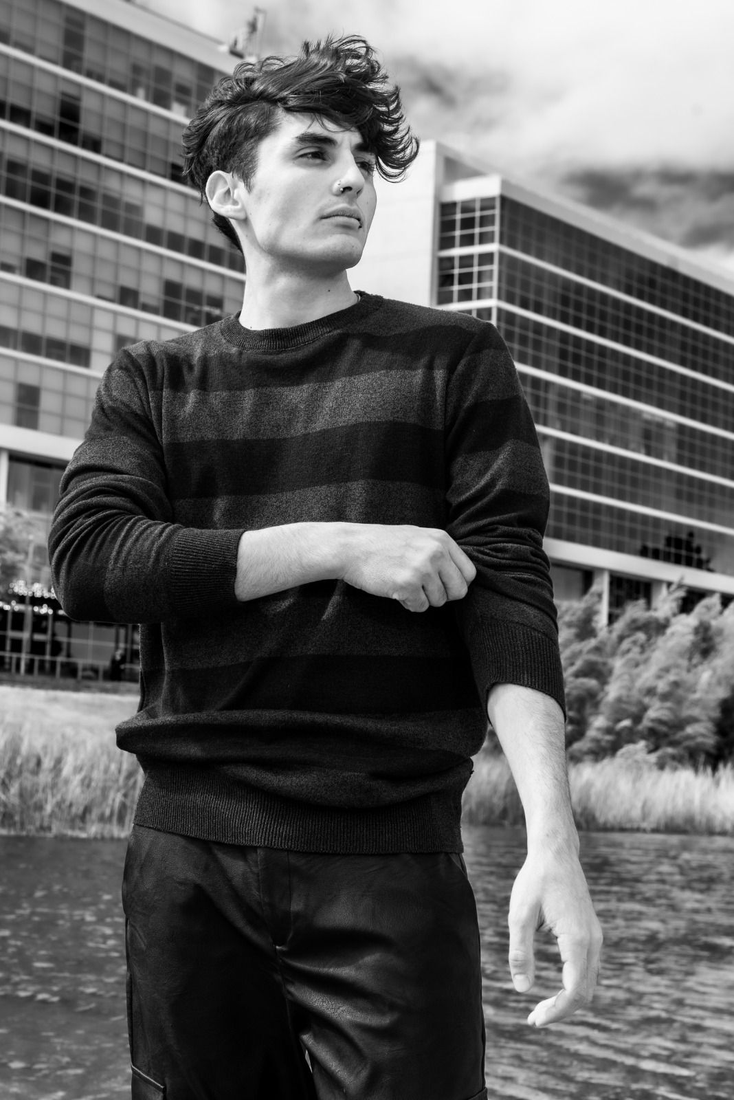 Hombre arremangándose, de pie cerca del agua con un edificio moderno al fondo.