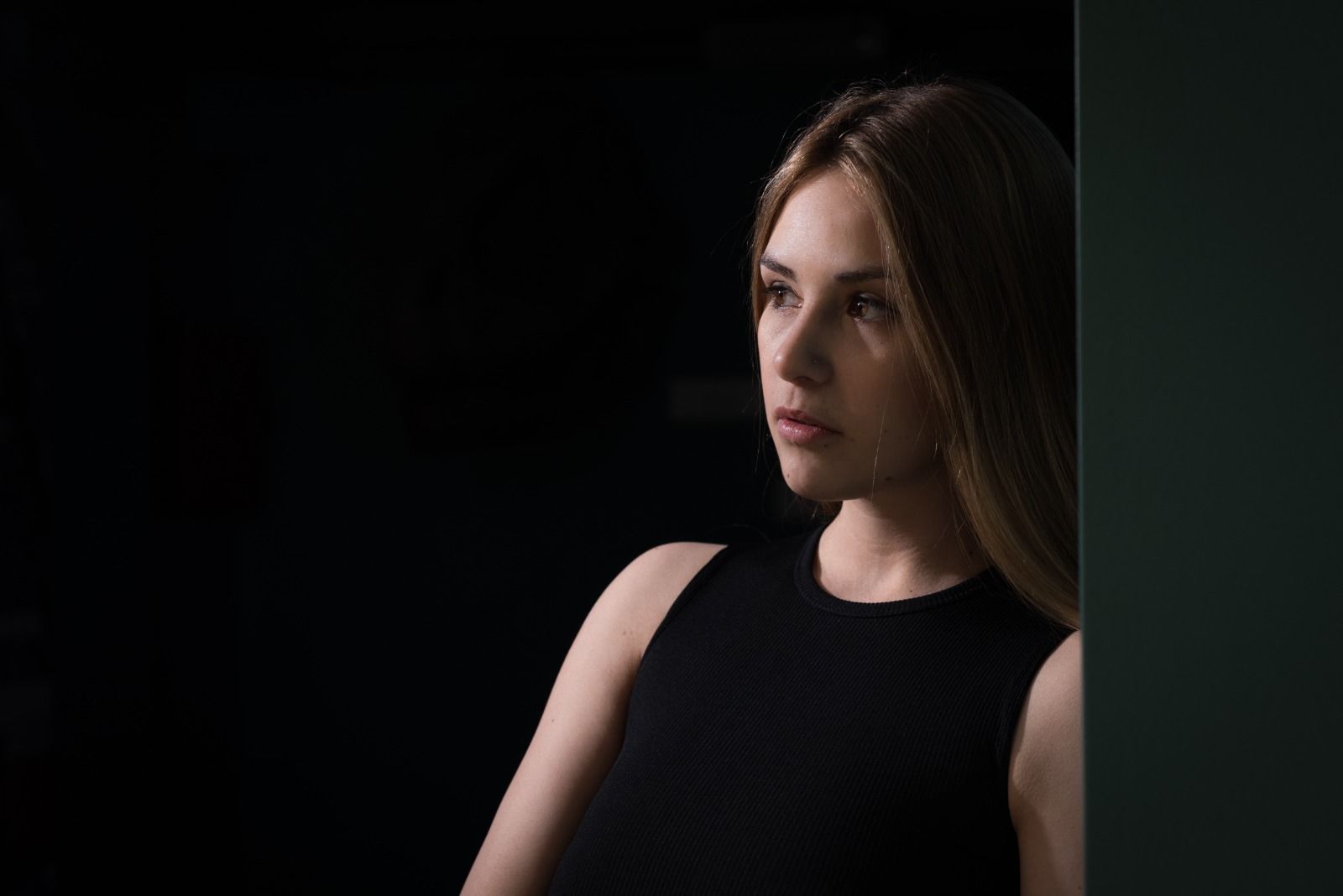 Mujer rubia con una blusa negra sin mangas, mirando hacia un lado. Apoyada sobre un fondo oscuro.