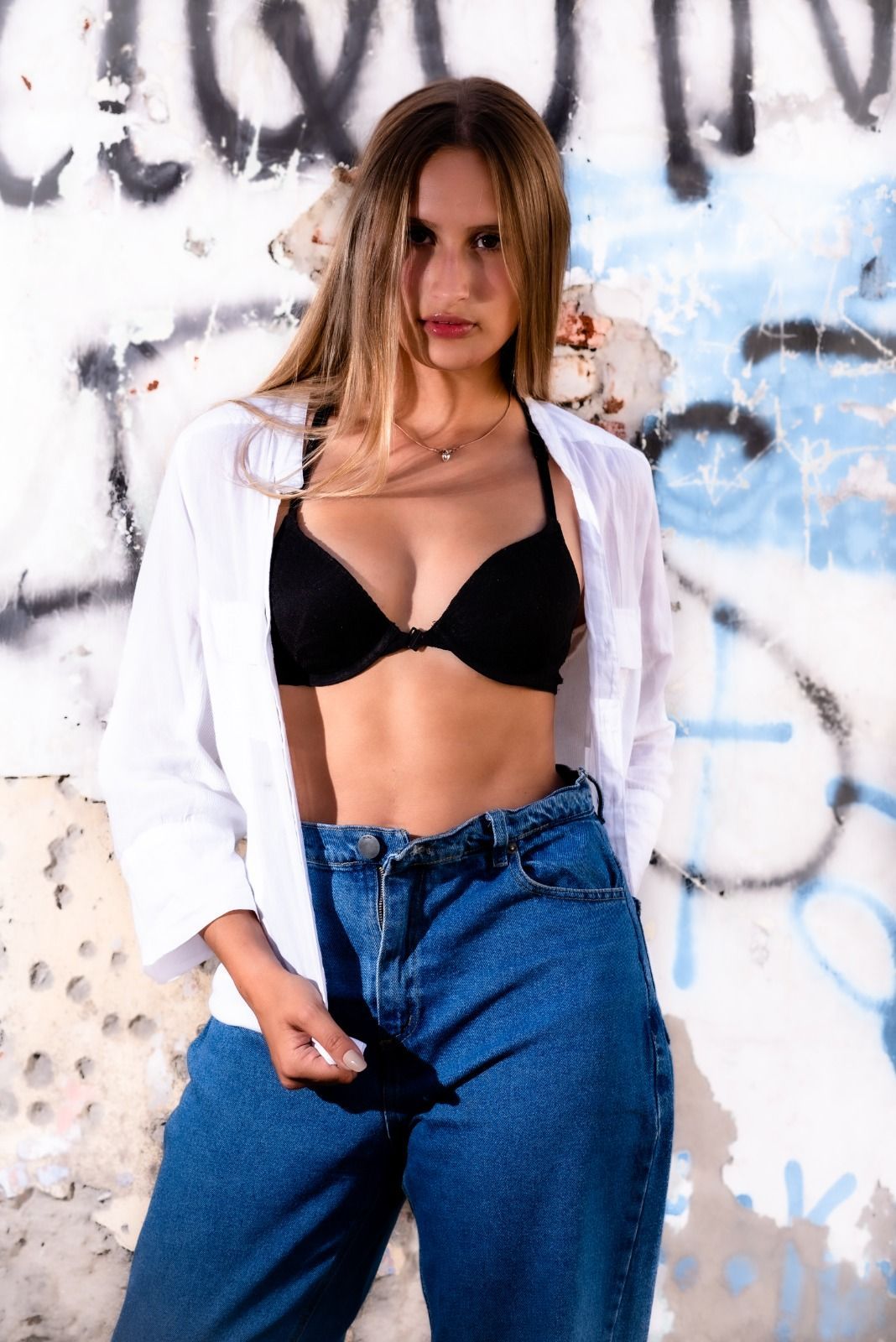Mujer con sujetador negro, camisa blanca y vaqueros posa frente a un muro de graffitis.