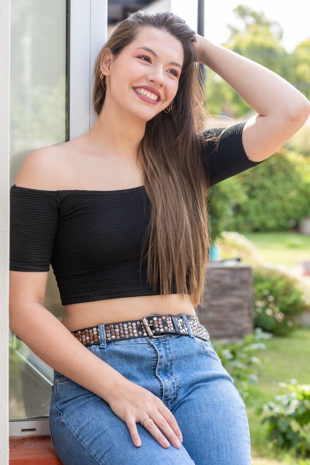 Una mujer con un top negro con hombros descubiertos y vaqueros sonríe, con la mano en el cabello, apoyada contra una ventana.