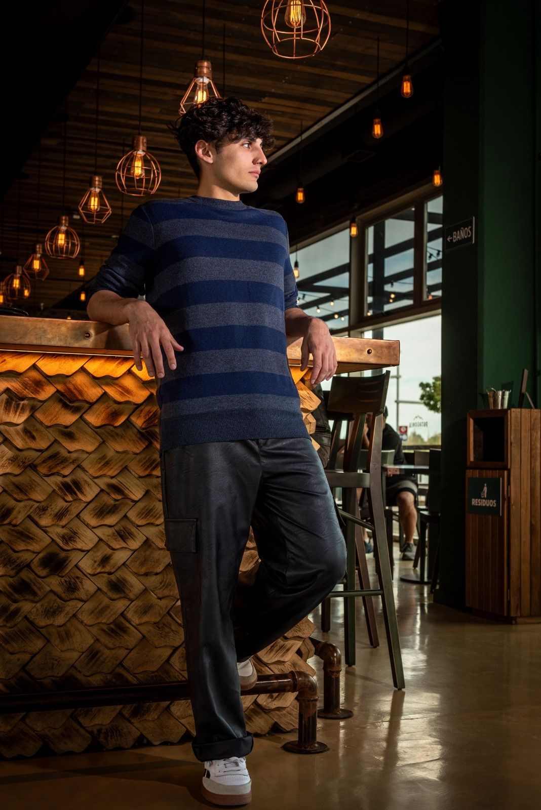 Hombre apoyado en una barra, vestido con camisa y pantalones a rayas