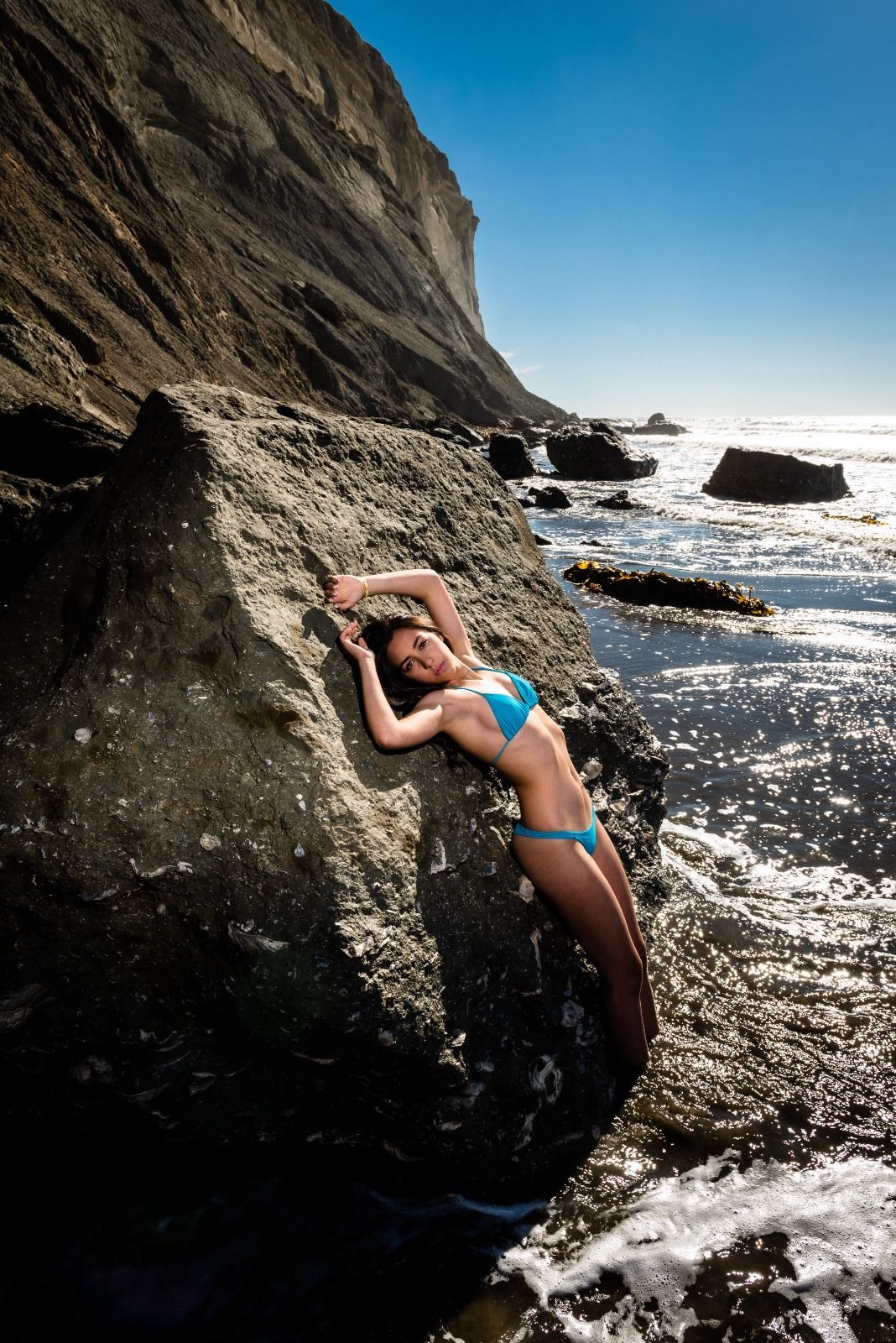 Mujer en bikini azul posa sobre una roca en la playa, cerca de un acantilado, en un día soleado.