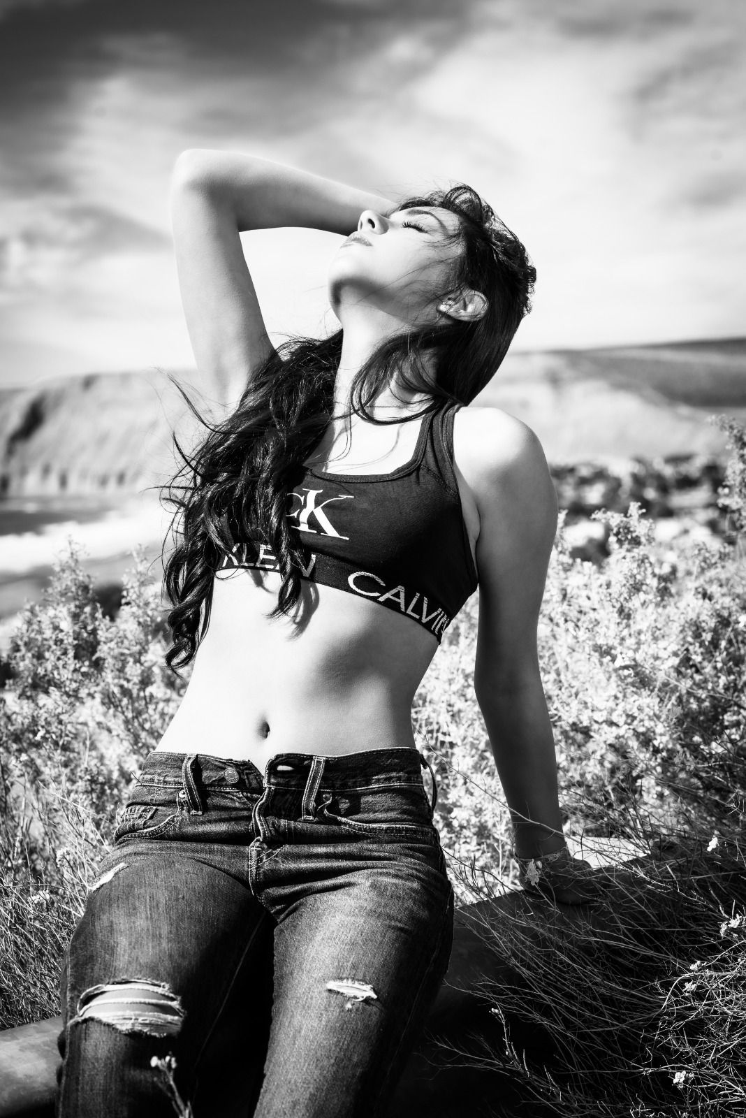 Mujer en sujetador deportivo y vaqueros, recostada al sol, al aire libre.