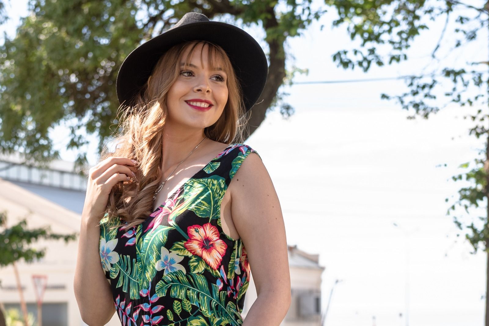 Una mujer con un vestido floral y un sombrero negro sonríe, mirando hacia un lado, afuera, cerca de los árboles.