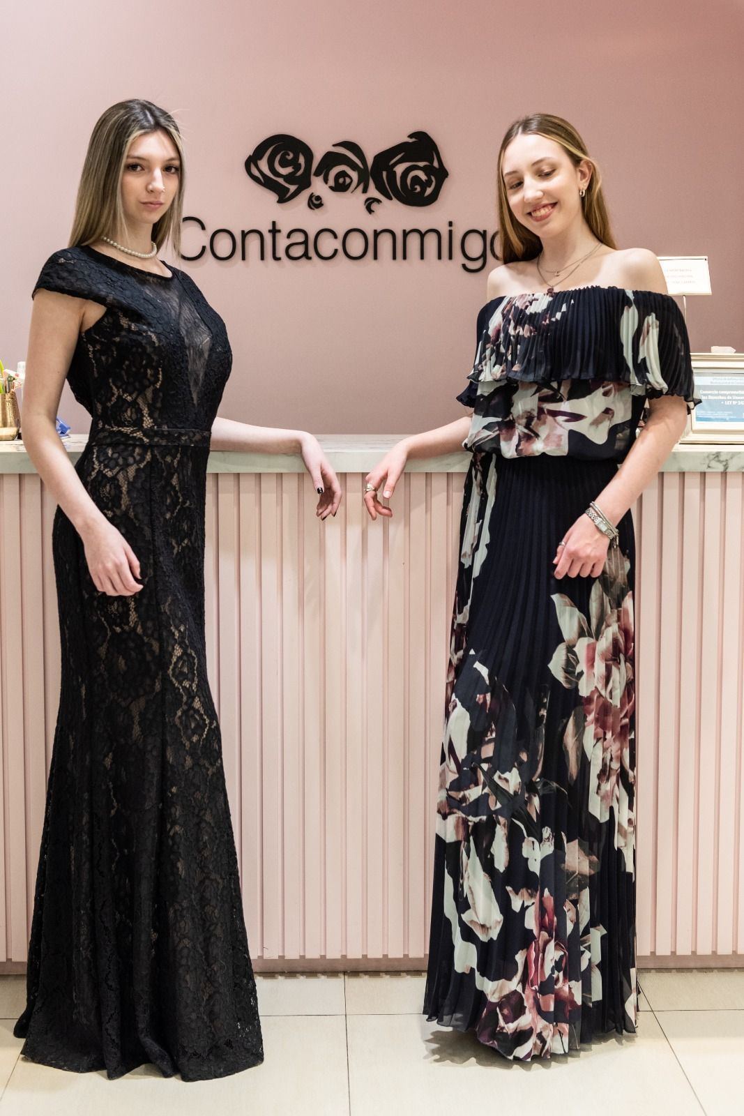 Dos mujeres con vestidos se apoyan en un mostrador rosa.