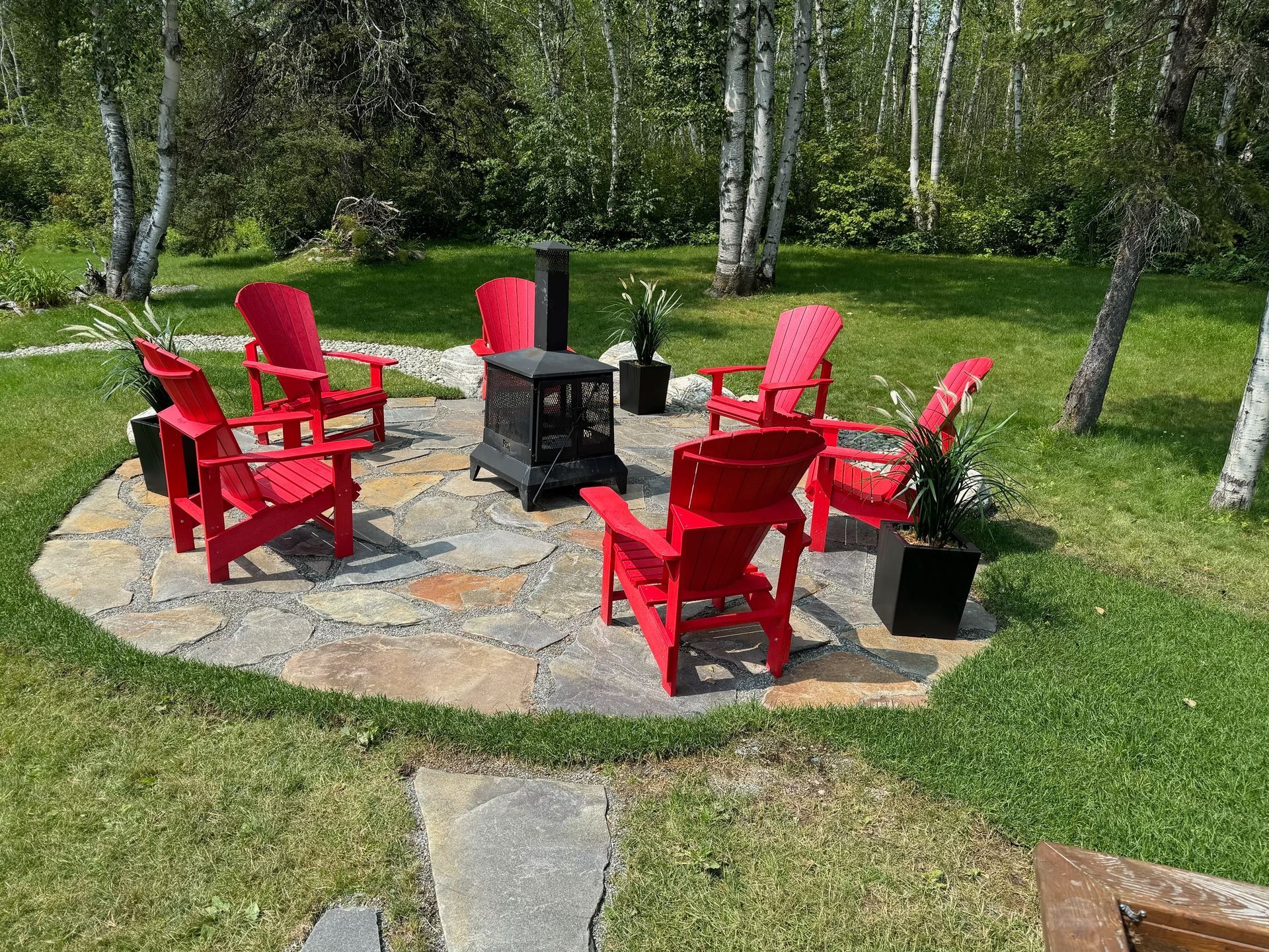 Des chaises Adirondack rouges entourent un foyer sur une terrasse en pierre dans une cour gazonnée.