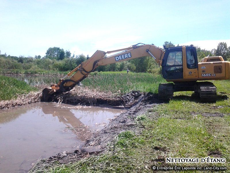 Une excavatrice jaune nettoie un étang dans un champ.