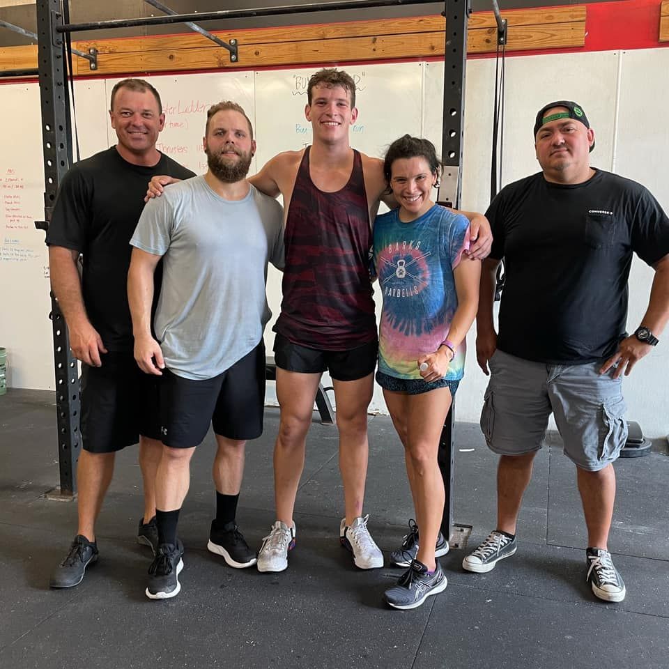Five people posing in a gym; diverse body types and clothing. Smiling, arms around each other, standing in front of a pull-up bar.