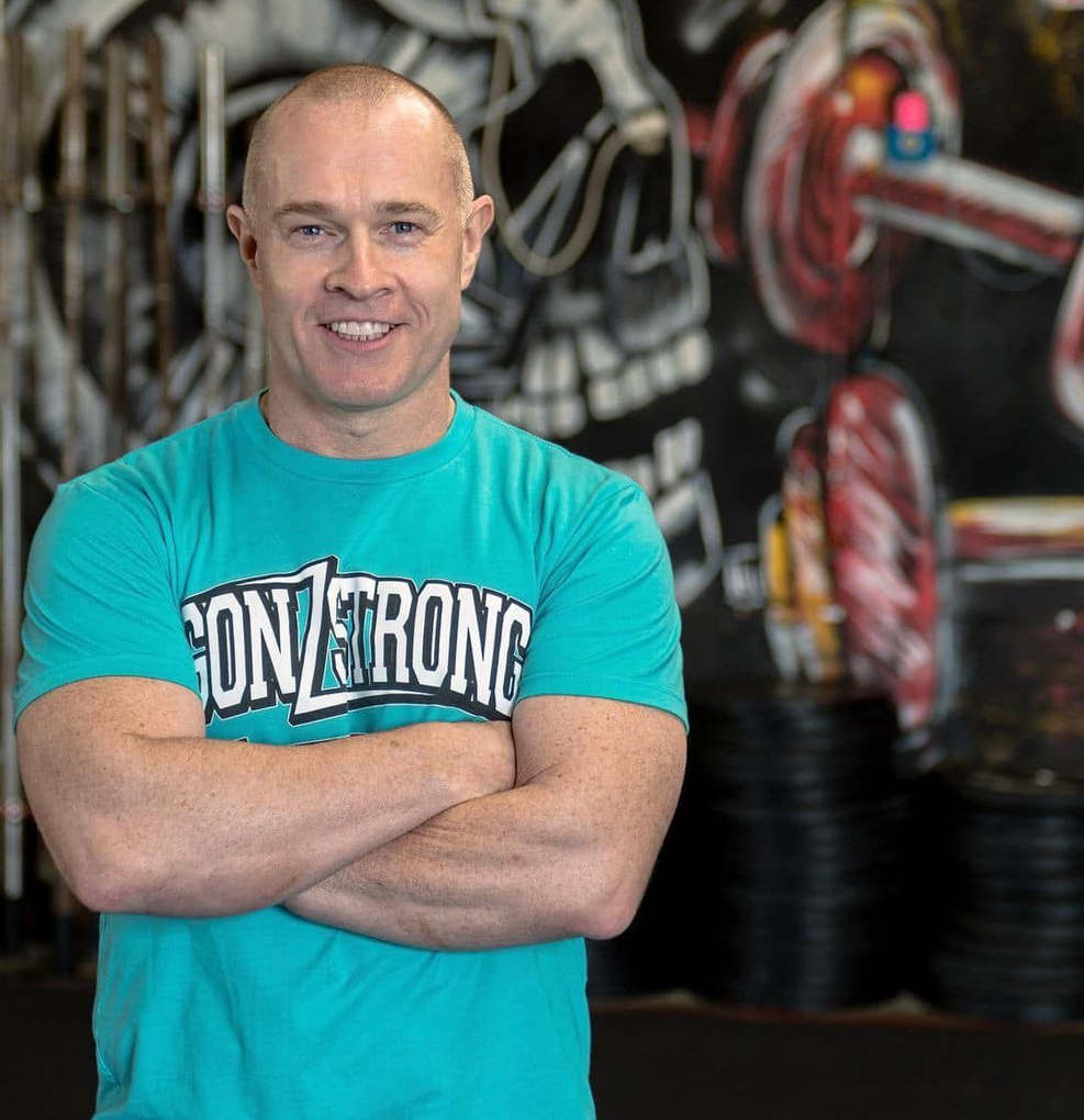 Man in teal t-shirt with crossed arms smiles at the viewer in a weightlifting gym.