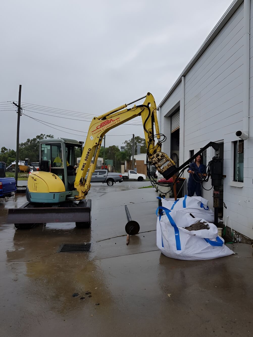 A Yellow Excavator is Parked in Front of a White Building — Q-Pile Foundations in Coolum Beach, QLD