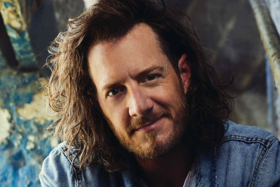 Tyler Hubbard. with long hair and a beard is standing in front of a blue wall.