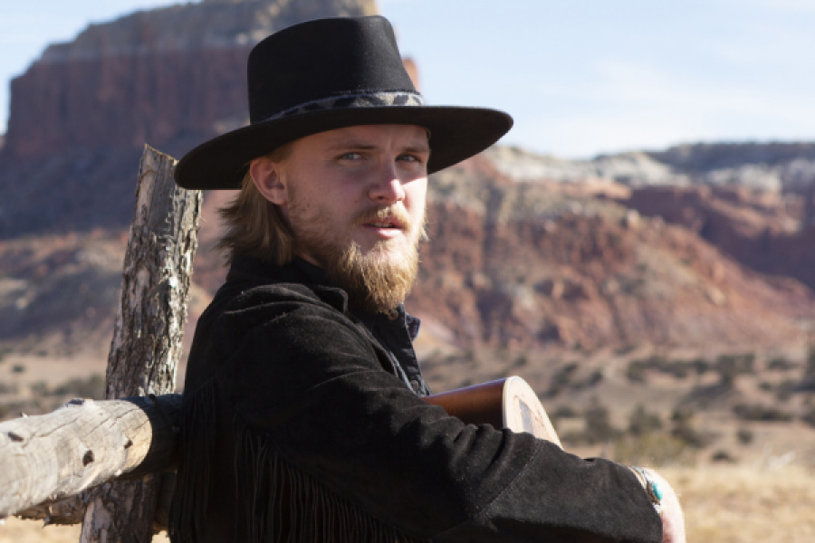 A man in a black hat leans against a wooden fence post. Jackson+Dean