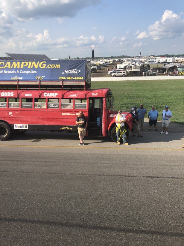 A group of people are standing in front of a red bus that says camping.com
