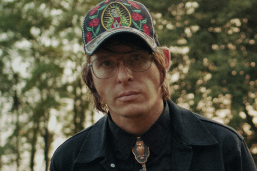 A man wearing a hat and glasses is standing in front of trees.