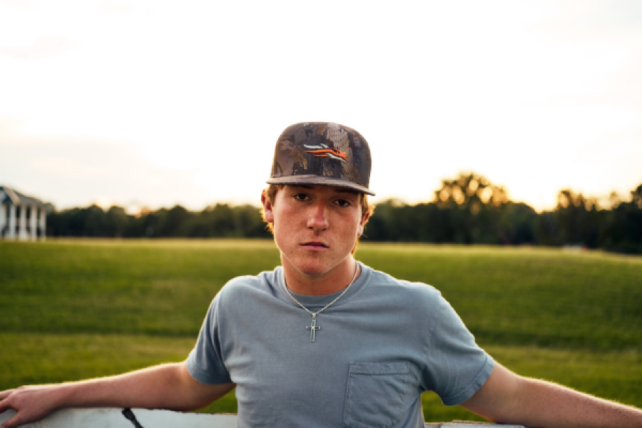 A young man wearing a baseball cap and a necklace is leaning on a fence.