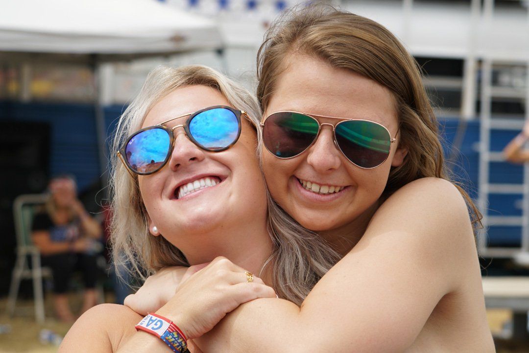 Two women wearing sunglasses are hugging each other and smiling.