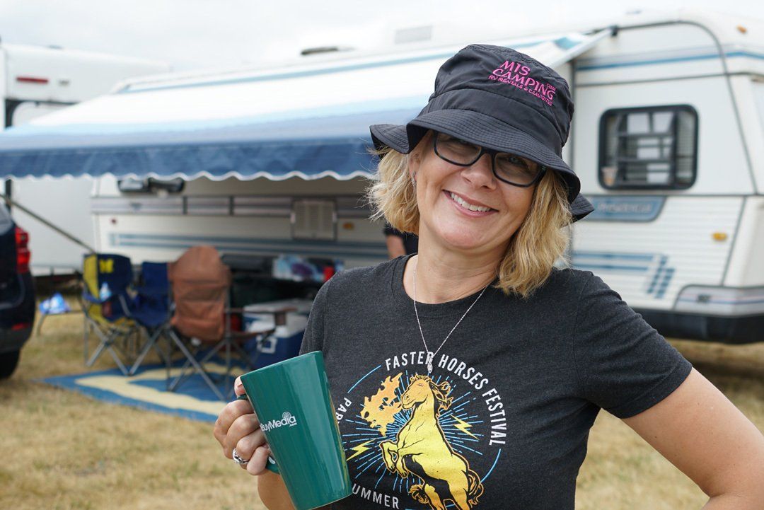 A woman wearing a hat and glasses is holding a cup in front of a trailer.