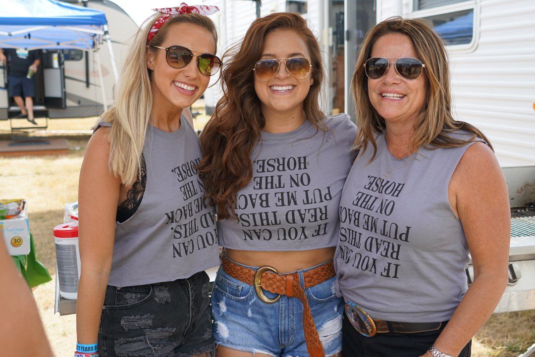 Three women are posing for a picture and one of them is wearing a shirt that says put me back on the horse