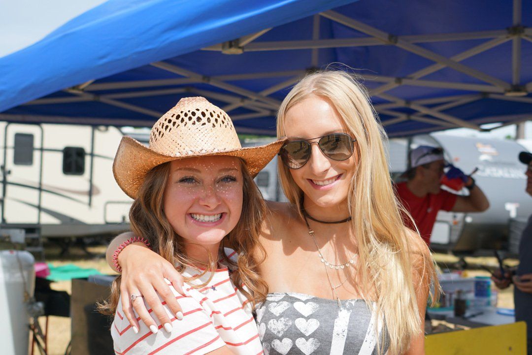 Two women are posing for a picture under a blue tent . one of the women is wearing a cowboy hat.