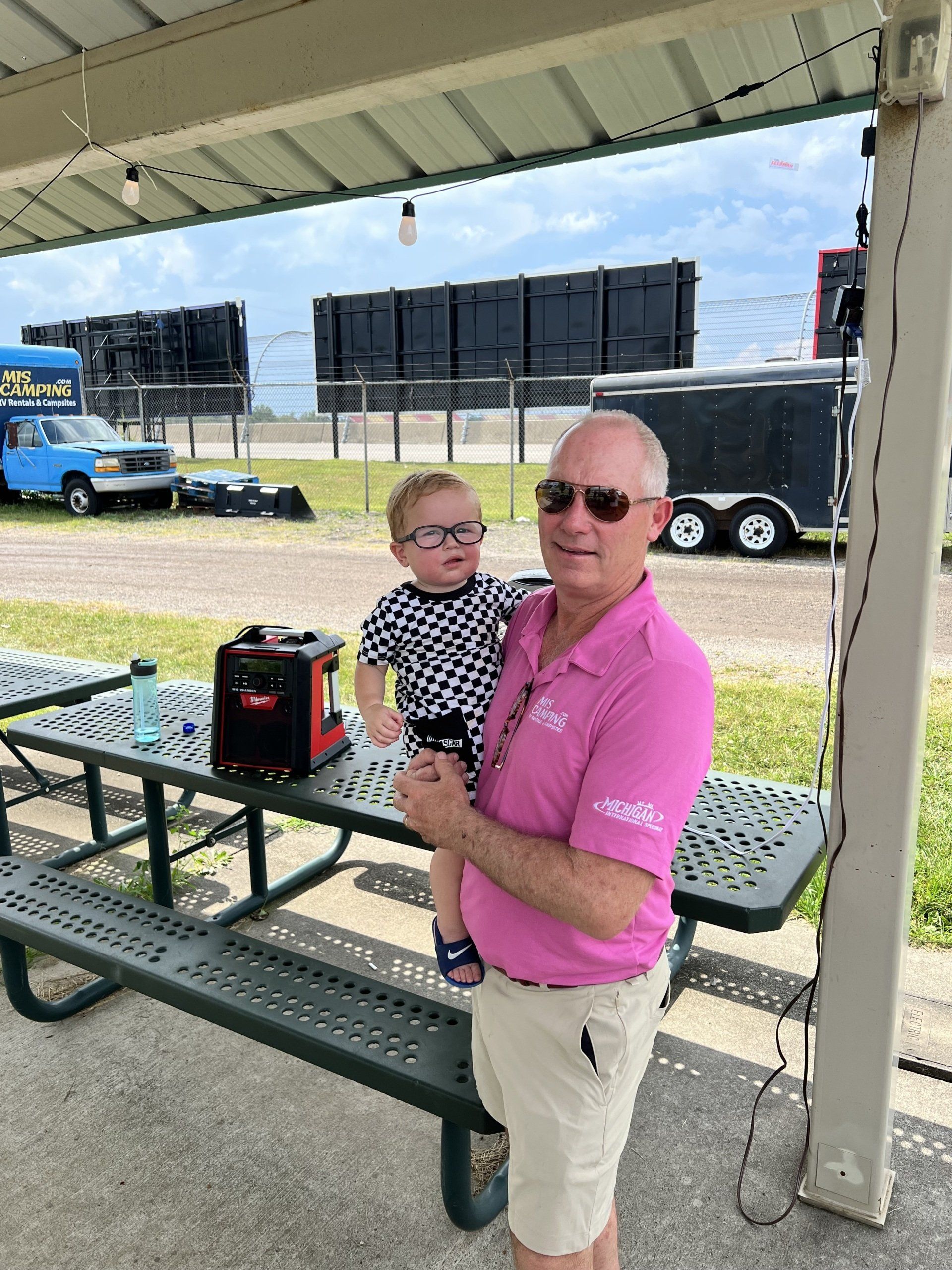 A man in a pink shirt is holding a baby in front of a picnic table.