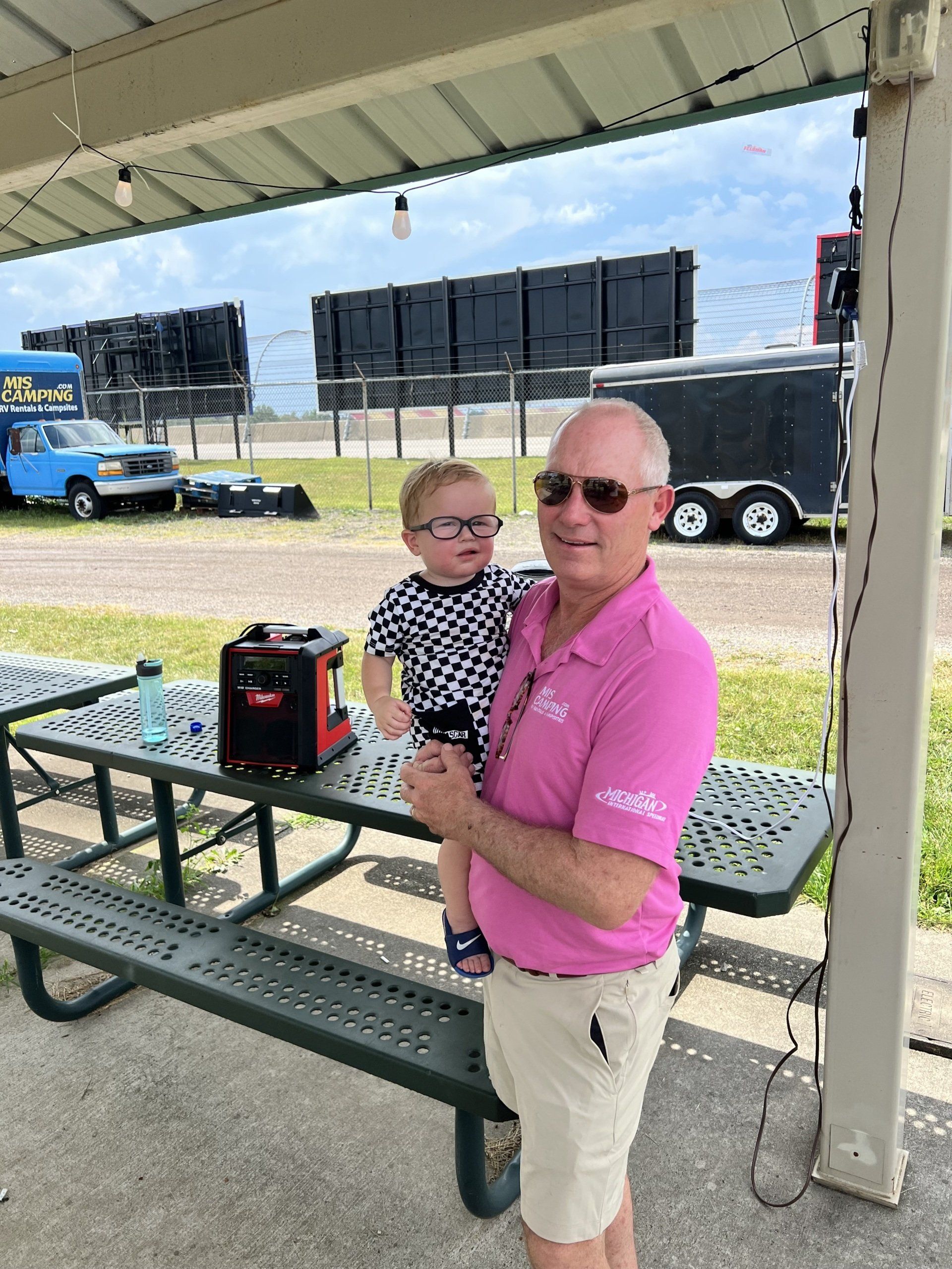 A man in a pink shirt is holding a baby in front of a picnic table.