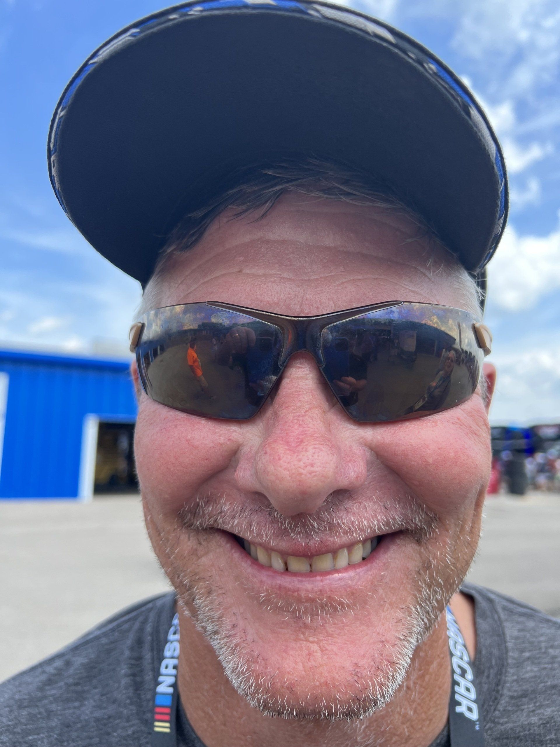 A man wearing sunglasses and a nascar lanyard smiles for the camera