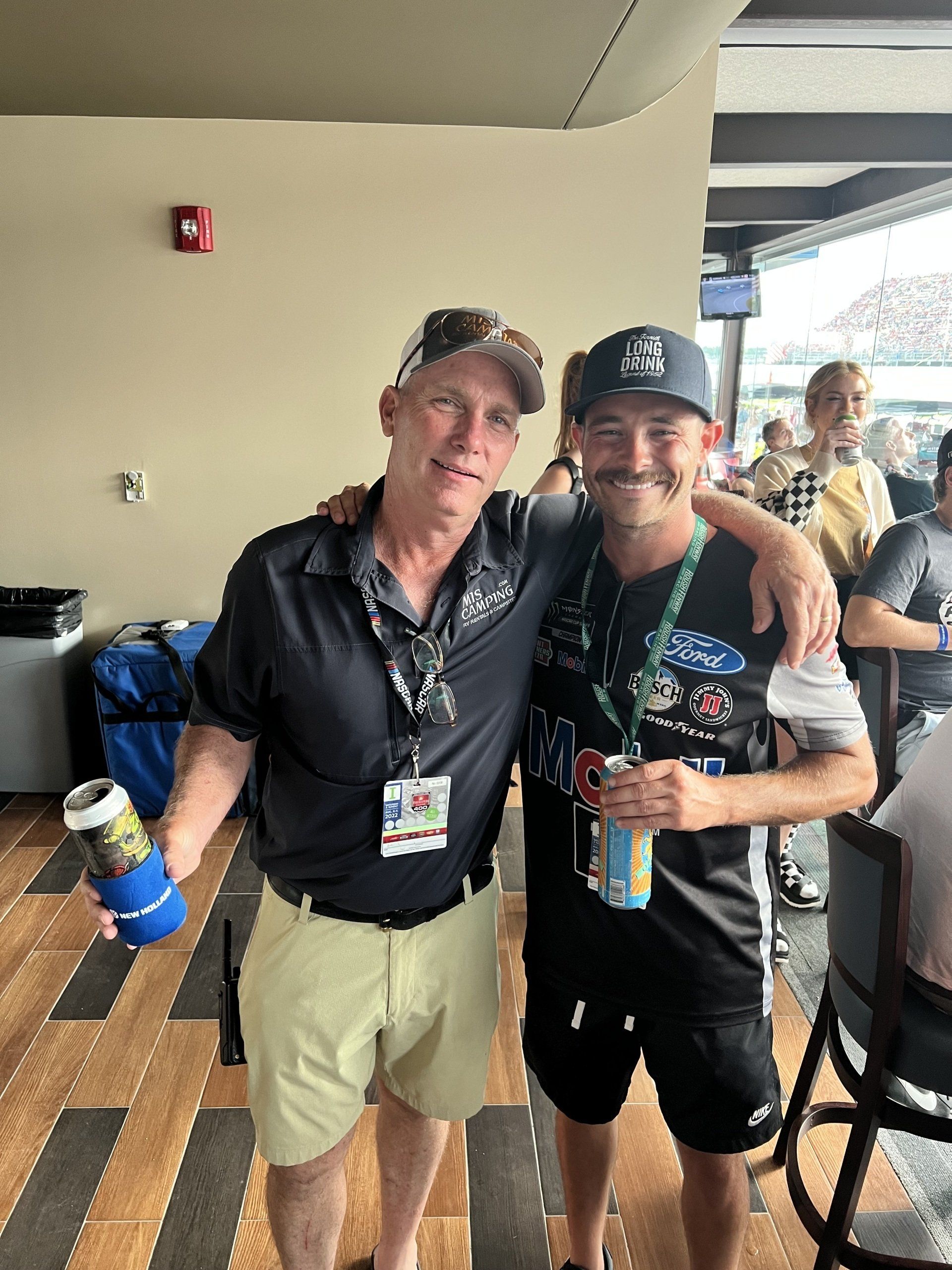 Two men are standing next to each other in a room holding cans of beer.