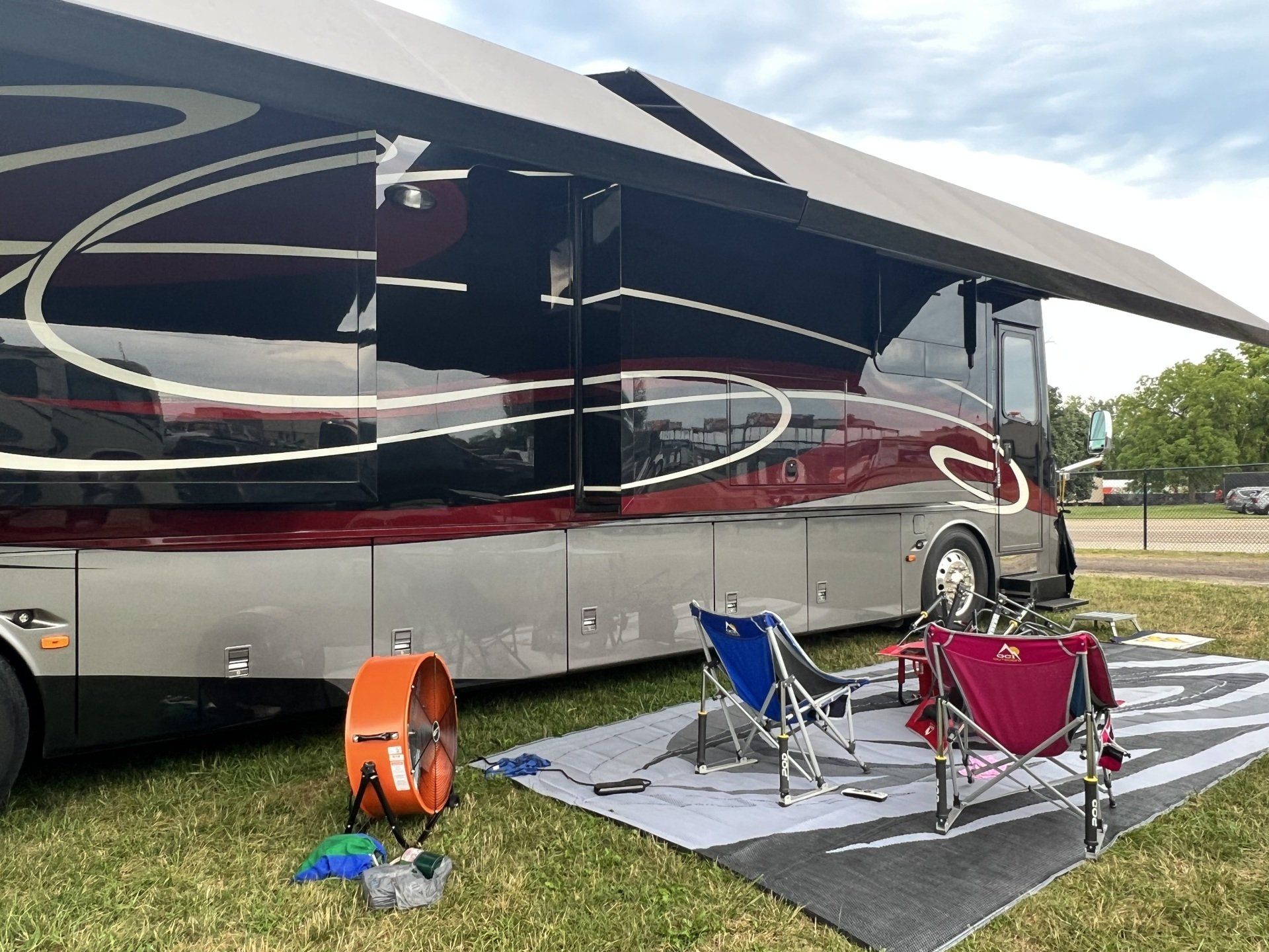 A large rv is parked in a grassy field with chairs and a fan.